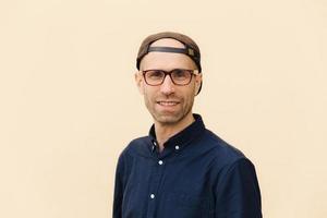 Portrait of satisfied Caucasian male with stubble, has gentle smile, wears stylish cap and shirt, looks through spectacles, poses against beige background. Hipster guy being in good mood. Horizontal photo