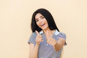 Shot of attractive European female gives double thumb up, has positive expression, has white teeth with brackets, dressed casually, makes okay and approval gesture. People and body language. photo