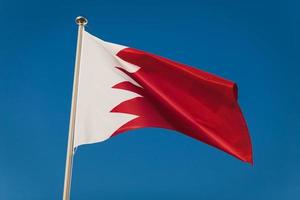 bandera roja y blanca de bahrein, capital manama. bandera nacional en el asta de la bandera frente al cielo azul. textil ondulante. vista de cerca foto