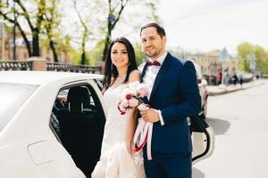 Lovely married couple stand next to each other near car, going in restaurant to celebrate their wedding, have party, invite friends and relatives. Newlyweds pose outdoor after registration of marriage photo