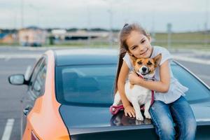 Small attractive female child embraces her favourite dog, sit together at trunk of car, have rest after stroll, enjoy summer day, have friendly relationship. Children, pets and lifestyle concept. photo