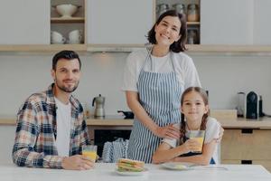 Positive friendly father, mother and their small daughter being in good mood, spend time together, pose at kitchen, gather to have breakfast, enjoy domestic atmosphere and day off. Time to eat photo