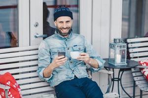 retrato de un hombre barbudo con una amplia sonrisa, se sienta en un banco, usa un teléfono celular moderno, navega por las redes sociales, feliz de recibir buenas noticias, bebe bebidas aromáticas. gente, tecnología, concepto de estilo de vida foto