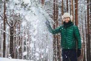 Portrait of cheerful handsome male wears fur cap with ear flap and yellow and green jacket, throws snow in air, enjoys free time spending in beautiful winter forest. People, relaxation concept photo