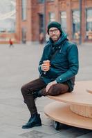 Concept of drinks and lifestyle. Carefree satisfied European man with bristle enjoys sunny day and hot beverage, smiles happily, sits against street background. Outdoor shot of handsome hipster photo