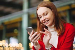 Positive pleased female model uses mobile phone data, uses chat application, holds modern cell phone, smiles positively, dressed in red coat, waits for answer or call from boyfriend, has brown hair