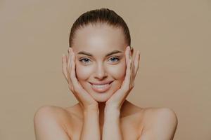 Skin care and beauty concept. Headshot of beautiful cheerful woman touches face gently, has perfect smile, healthy skin after cleaning or applying facial mask, isolated over beige background photo