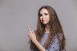 Shot of serious pleasant looking brunette female with blue eyes, dressed casually, points with index finger at blank copy space, isolated over grey background. Woman advertizes new product indoor photo