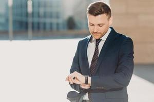 Handsome attractive male checks time, hurries for business meeting, looks at watch, dressed in formal black suit, poses outdoor near office building. People, business and occupation concept. photo