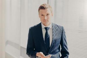 Indoor shot of clever businessman in formal clothes, holds smart phone, prepares for business meeting in office, has work break, stands near window with shutter. Technology and occupation concept photo