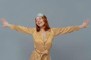 Positive relaxed woman wears blindfold and striped robe stretches arms sideways, smiles broadly, closes eyes, isolated over grey background, wakes up in bedroom, slept well. Good morning concept photo