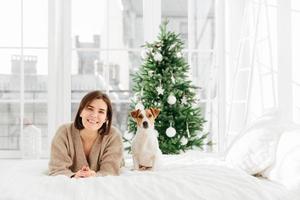 Photo of happy young woman and pedigree dog wait together for New Year, pose at home in bedroom with white walls, big windows, enjoy comfort on bed, Christmas tree in background, smile at camera