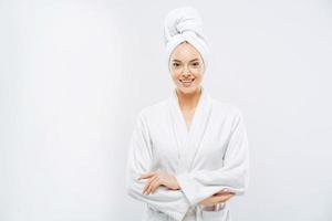 Beautiful smiling young woman has soft healthy skin after taking shower, wears bath robe and towel wrapped on head, enjoys spare time at home, isolated over white background. Wellness concept. photo