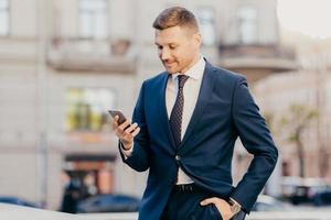 Photo of successful male entrepreneur holds modern smart phone in hands, reads news from financial exchange, browses internet, makes online payments, has work break, going to cafe or restaurant