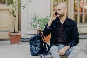 Handsome bald male with beard, has rest after long stroll outdoor, speaks via cell phone, focused aside, rucksack near him, poses against builduing background. People and communication concept photo