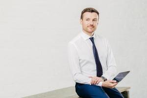 Thoughtful young male economist dressed in elegant luxury clothes, works on touch pad during break, poses in modern office corridor, thinks about cooperation, isolated over white background. photo