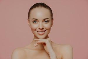 Pretty young European woman touches chin gently, enjoys flawless of skin after beauty procedures, poses nude, has natural makeup, isolated on pink background. Beauty, spa and wellness concept photo