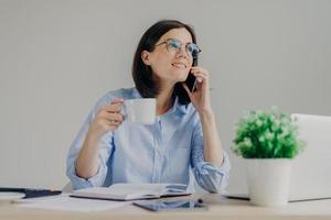 Gorgeous pleased businesswoman has cell phone conversation while has coffe break after working on notebook, shares her success with close person, isolated over grey background. Occupation concept photo