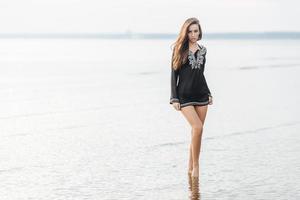 Horizontal shot of pretty female in black blouse, demonstrates her perfect body and slender legs, stands against ocean background, looks with confident expression at camera, poses for magazine photo