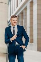 Vertical shot of handsome male with appealing appearance, dressed in formal black suit, keeps hand in pocket, waits for business partners, going to have negotiations, has positive expression. photo