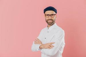 Positive male entrepreneur with self confident satisfied facial expression, keeps hands crossed, stands in profile, looks directly at camera, poses indoor against pink background with free space photo