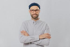 una foto horizontal de un diseñador masculino satisfecho y seguro de sí mismo usa un elegante sombrero, vestido con una camisa blanca, mantiene los brazos cruzados, tiene un rastrojo oscuro, aislado sobre fondo blanco. concepto de personas y estilo