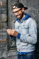 Vertical portrait of hipster guy in denim shirt, cap and eyewear holding modern telephone in his hands having glad expression while communicating with his girlfriend. Lifestyle and technolgy concept photo