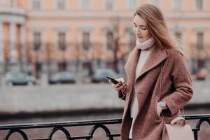 Beautiful woman in warm coat, holds modern cell phone, messages in social networks, connected to free wifi, walk in city, street style, blurred background with copy space for your advertisement photo