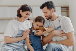 Affectionate little girl embraces pedigree dog with love, mother and father pose near, spend free time at kitchen, prepare delicious breakfast. Happy family and their pet love each other very much photo