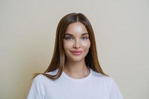 Tender feminine woman looks with admiration and affection, wears white t shirt, has makeup, healthy skin, dark hair, looks at something wonderful, isolated over beige studio wall. Beauty, cosmetology photo