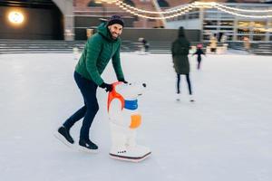 Delightful male skates on skate ring with help of special aid, practices to make some tricks, has happy expression as being on ice for first time, has fun with friends. People and leisure concept photo