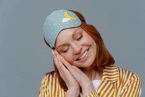 Close up shot of lovely tender woman has ginger hair, minimal makeup, leans at palms pressed together, smiles positively, wears blindfold on head, sees pleasant dreams. Healthy lifestyle concept photo