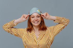 Positive redhaired young female wears eye mask for sleep on head, smiles gladfully, dressed in home wear, has sweet dreams at night, isolated over grey background, enjoys peaceful atmosphere photo