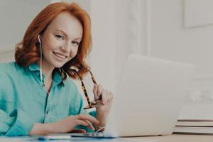 Smiling good looking female employee talks on video call, has web conference with colleagues, watches webinar on modern laptop, poses in coworking space. Student learns foreign languages online photo