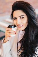 Beauty, youth, lifestyle and relaxation concept. Young brunette female with well-shaped thin eyebrows, healthy skin and red manicure holding takeaway coffee looking pleasantly into camera smiling photo