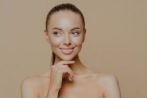 Smiling well groomed young woman with beauty face care, healthy glowing skin, touches chin gently and looks aside, poses shirtless against brown studio background, loves her skin, feels refreshed photo