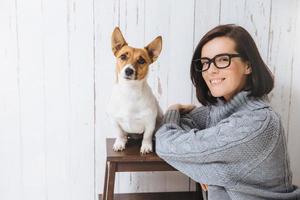 foto de una adorable mujer contenta y satisfecha que usa anteojos y un suéter suelto y cálido, se para cerca de su perro favorito que se sienta en la silla, mira la cámara y va a caminar al aire libre. personas y mascotas.