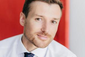 Close up portrait of attractive unshaven male looks at camera, involved in business affairs, being confident in his strength, poses against blurred red and white wall. Man boss with bristle. photo