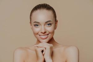 Close up shot of adult woman with fresh daily makeup, smiles toothily and keeps hands under chin, stands half naked indoor, feels refreshed after cosmetic procedures. Beauty and face care concept photo