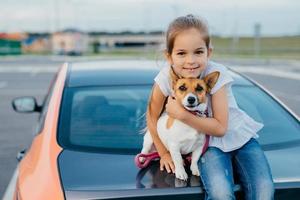 Little girl with appealing appearance, hugs her favourite pet, have journey together with parents by car, sit at trunk, pose for making photo. Children, animals, rest and transportation concept photo