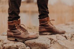 Cropped shot of unrecognizable man wears old shoes, has walk outdoor, blurred background. Boots on stones. Fashion concept photo