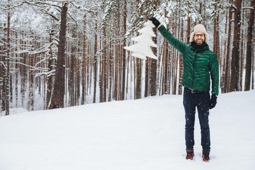 toma horizontal de buen aspecto de un hombre barbudo con expresión feliz, demuestra un abeto artificial que compró, camina sobre un clima nevado en un hermoso bosque. concepto de felicidad foto
