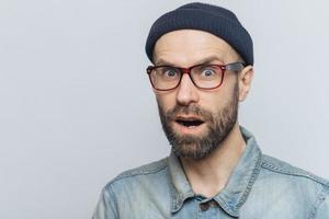 Suprised terrified unshaven male stares at camera and keeps mouth widely opened, wears fashionable clothing, expresses great shock as hears unexpected news from interlocutor, isolated on grey wall photo