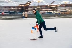 Young male begginer learns skating with help of special figure, leans at it, being on ice rink covered with snow, has cheerful expression, poses in camera. Man uses special equipment for skating photo