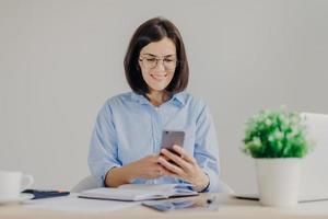 Happy female employee works on developing new project in office, searches information on websites via modern electronic gadgets, uses free internet connection in her cabinet, has happy expression photo