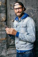 Fashionable young bearded man in cap, denim shirt and eyeglasses holding smartphone in his hands making photos while standing near fissure surface. Happy hipster guy with gadget in his hands