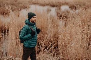 toma lateral de un hombre activo vestido con una chaqueta y un sombrero abrigados, lleva una mochila, camina en una zona rural, posa cerca del fondo del lago, disfruta del aire fresco, un ambiente tranquilo. concepto de personas y estilo de vida foto