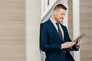 Prosperous businessman in formal clothes reads notification on digital tablet, installs new application, connected to wireless internet, poses indoor. People, business and modern technology concept photo