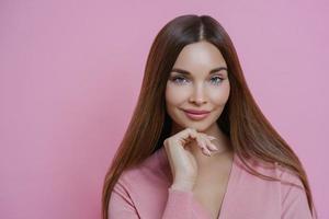 Pleasant looking brunette female fashion model keeps hand under chin, has full lips and applealing appearance, wears rosy jumper in one tone with background. Women, cosmetic and skin care concept photo