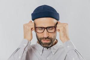 Close up shot of stressful unshaven man keeps hand near temples, has terrible headache, suffers from pain, wears headgear, spectacles, isolated over white background. Exhausted dissatisfied male photo